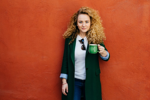 Mooie zekere blonde krullende vrouw elegant jasje dragen en jeans die groene mok met thee houden.