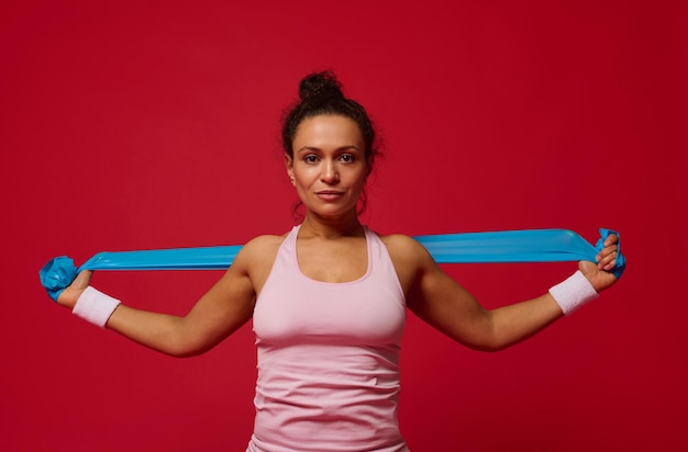 Mooie zelfverzekerde sportvrouw die traint met een elastische band op een rode achtergrond en naar de camera kijkt Kopieer advertentieruimte