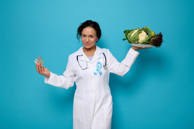 Mooie zelfverzekerde vrouwelijke arts in witte medische jurk met blauw lint, symbool van World Diabetes Awareness Day, houdt een bord met gezond veganistisch eten en blister met medicijnen in handen