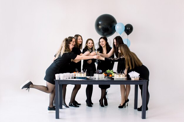 Mooie zeven meisjes in zwarte jurken die de martini-glazen met elkaar aanraken, verjaardag vieren, bij de gedecoreerde tafel staan met cake, drankjes, desserts en met ballonnen