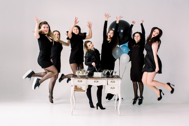 Mooie zeven meisjes in zwarte jurken, verjaardag of vrijgezellenfeest vieren, staan en springen bij de gedecoreerde tafel met cake, drankjes, desserts en met ballonnen