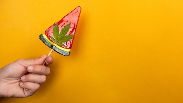 Mooie zoete rode lolly met marihuana in de hand op gele achtergrond, snoep met cannabis.