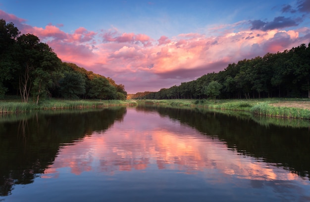 Mooie zomer zonsondergang aan de rivier met blauwe lucht, rode en oranje wolken, groene bomen en water met reflectie.
