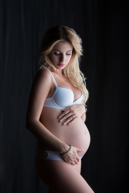 Mooie zwangere blonde vrouw staat en knuffelt haar buik op een zwarte muur.