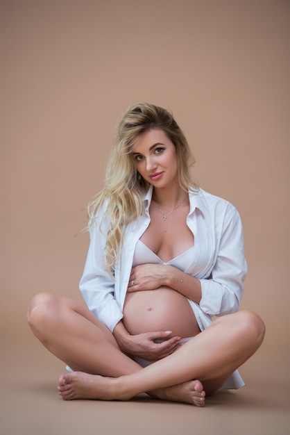 mooie zwangere blonde vrouw zit in een wit overhemd en ondergoed op een beige muur