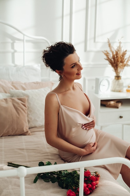 Mooie zwangere dame die vredig kijkt terwijl ze op het bed zit naast een boeket rozen en haar buik streelt