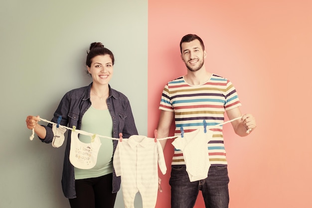 Mooie zwangere vrouw en haar man verwachten een baby met baby bodysuits en glimlachen op een kleurrijke achtergrond