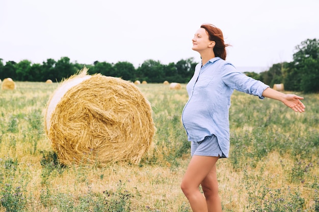 Mooie zwangere vrouw in de natuur buitenshuis Vrouw met opgeheven armen op achtergrond van tarweveld met hooibergen op zomerdag Foto van zwangerschap moederschap verwachting moeder wachten van een baby