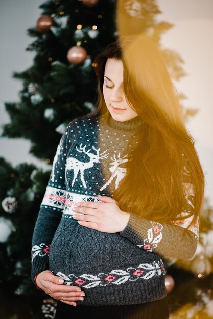 Mooie zwangere vrouw in gezellige kleding in de buurt van boom Gelukkig Nieuwjaar en vrolijk kerstfeest Kerst versierd interieur Zwangerschap vakantie mensen en verwachting concept Close-up