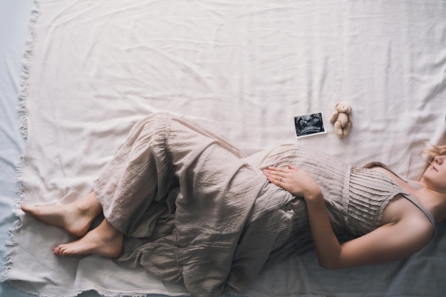 Mooie zwangere vrouw in jurk met handen op de buik liggend op de zijkant in profiel in bed verwachtende moeder die wacht en zich voorbereidt op de geboorte van de baby zwangerschapsfoto met een gelukkige en zachte stemming