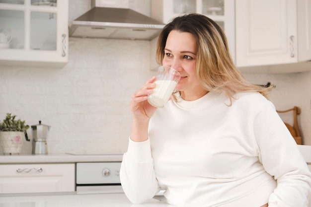 Mooie zwangere vrouw in wit pak drinkt melk in lichte keuken