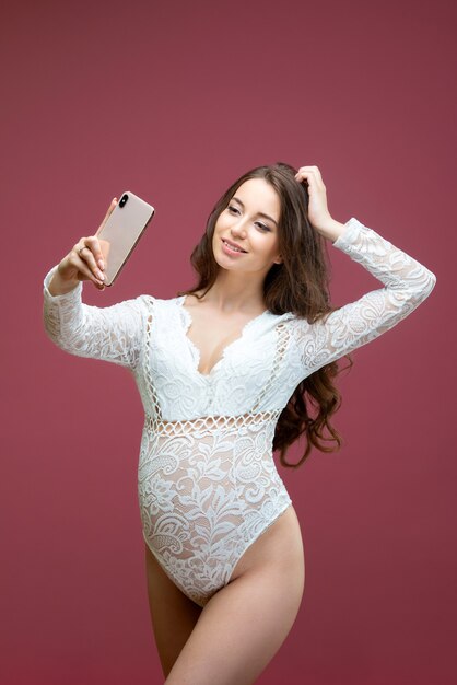 Mooie zwangere vrouw met lang haar gekleed in witte kanten lingerie met een pak doet een selfie op een roze achtergrond in de studio.