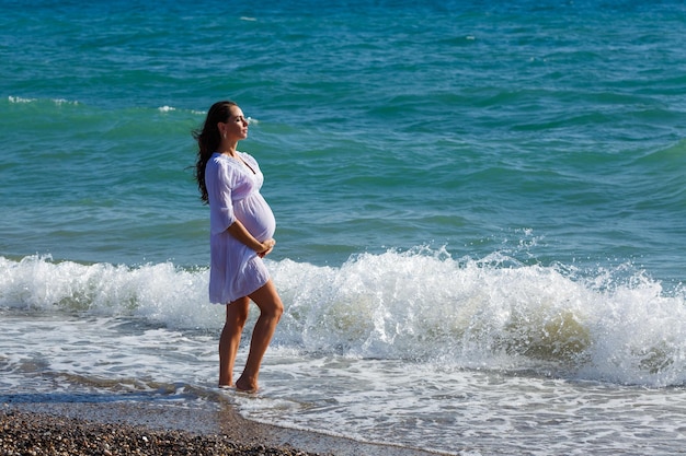 Mooie zwangere vrouw staande op het strand in lichte jurk in de wind Pleasure