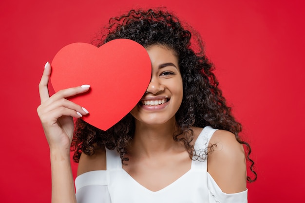 Mooie zwarte die oog behandelt met de valentijnskaartkaart van de hartvorm die op rood wordt geïsoleerd