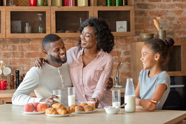 Mooie zwarte familie van drie die lunchen in de keuken, melk drinken en gebak eten, kopieer ruimte