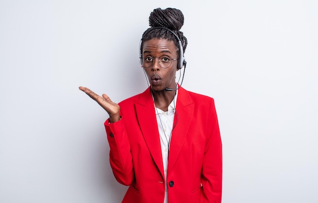 Mooie zwarte vrouw die verrast en geschokt kijkt, met open mond terwijl ze een object vasthoudt. telemarketing concept