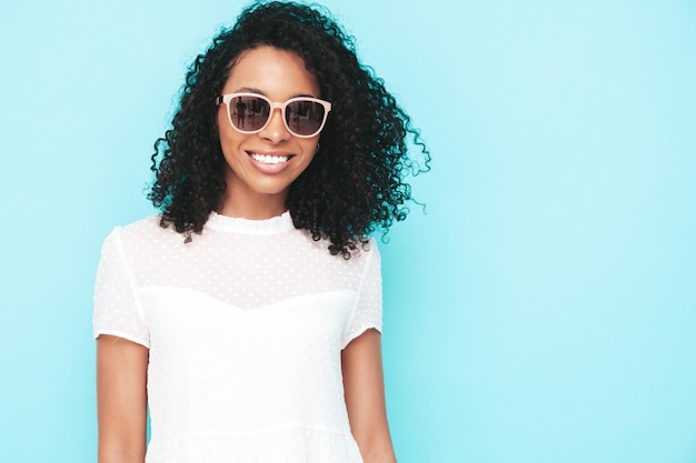 Mooie zwarte vrouw met afro krullen kapsel glimlachend model gekleed in witte zomerjurk Sexy zorgeloos vrouw poseren in de buurt van blauwe muur in studio gelooid en vrolijk In zonnebril geïsoleerd