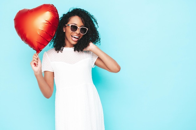 Mooie zwarte vrouw met afro krullen kapsel glimlachend model gekleed in witte zomerjurk sexy zorgeloze vrouw poseren in de buurt van blauwe muur in studio gebruind en vrolijk holding hart luchtballon