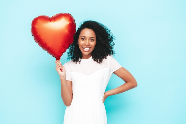 Mooie zwarte vrouw met afro krullen kapsel Glimlachend model gekleed in witte zomerjurk Sexy zorgeloze vrouw poseren in de buurt van blauwe muur in studio Gebruind en vrolijk Holding hart luchtballon