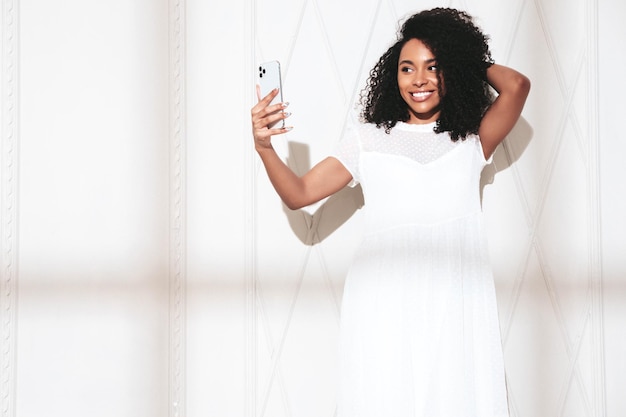 Mooie zwarte vrouw met afro krullen kapsel glimlachend model gekleed in witte zomerjurk Sexy zorgeloze vrouw poseren in de buurt van muur in studio op zonnige dag schaduw van raam Nemen selfie in brillen