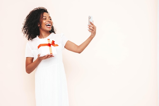 Mooie zwarte vrouw met afro krullen kapsel Glimlachend model gekleed in witte zomerjurk Sexy zorgeloze vrouw poseren in de buurt van witte muur in studio Gebruind en vrolijk Holding geschenkdoos Selfie nemen