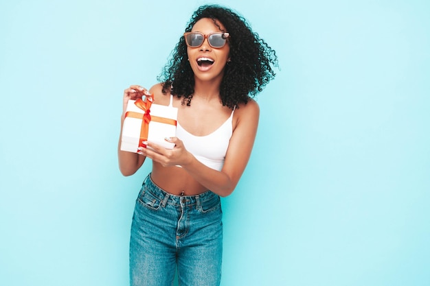 Mooie zwarte vrouw met afro krullen kapsel Glimlachend model gekleed in witte zomerkleren Sexy zorgeloze vrouw poseren in de buurt van blauwe muur in studio Gelooid en vrolijk Holding geschenkdoos geïsoleerd