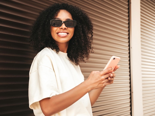 Mooie zwarte vrouw met afro krullen kapselLachende hipster model in witte tshirt Sexy zorgeloze vrouw poseren in de straat in de buurt van bruine muur in zonnebril kijken naar smartphone scherm met behulp van apps