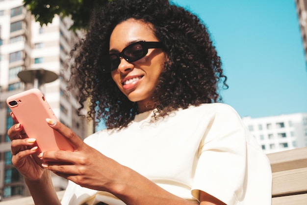 Mooie zwarte vrouw met afro krullen kapselLachende model in witte hoodie Sexy zorgeloze vrouw poseren op de straat achtergrond in zonnebril kijken naar smartphone scherm met behulp van apps