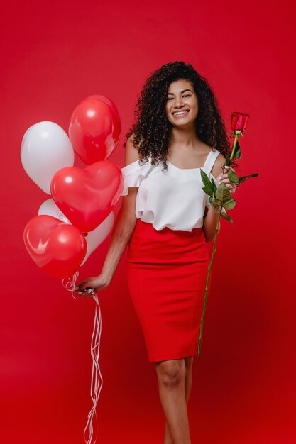 Mooie zwarte vrouw met roos en hartvormige ballonnen geïsoleerd op rood