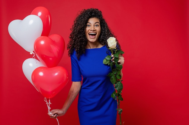 Mooie zwarte vrouw met witte roos en hartvormige kleurrijke ballonnen geïsoleerd op rode muur