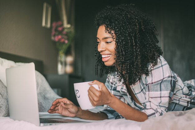 Mooie zwarte vrouw op bed met laptop en kopje koffie