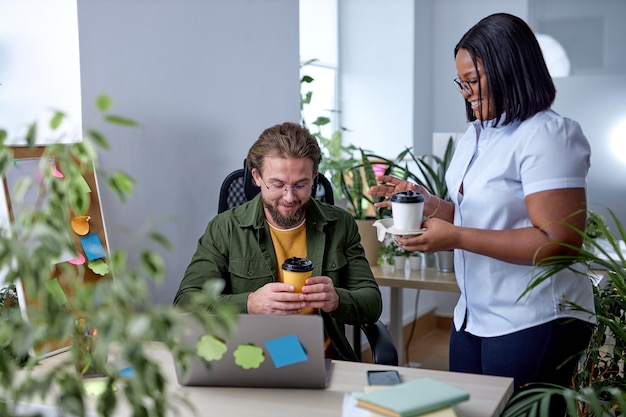 Foto mooie zwarte vrouwelijke manager in gesprek met collega-werknemer, met pauze met koffie. in het kantoor. jonge zwarte dame mentorcoach in gesprek met mannelijke collega die een gesprek heeft, glimlacht, leuk