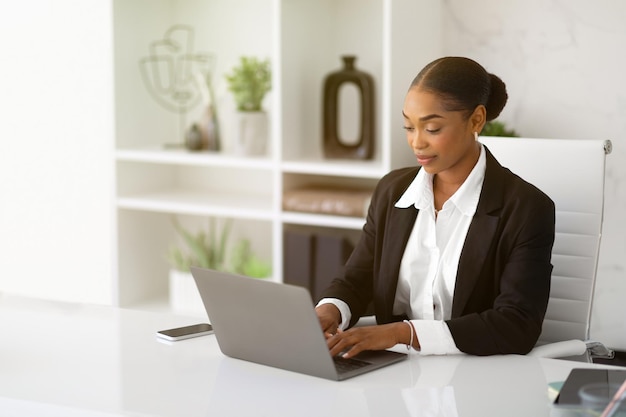 Mooie zwarte zakenvrouw in formele kleding die met een laptop werkt aan een bureau in het interieur van het kantoor