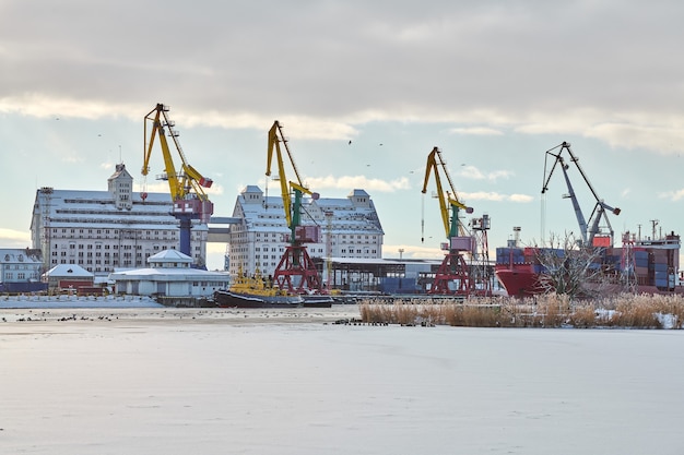 Пришвартованные грузовые суда и портовые краны в порту. Морской порт, грузовая контейнерная площадка, терминал контейнеровозов, верфь. Бизнес и коммерция, логистика. Зимняя индустриальная сцена.