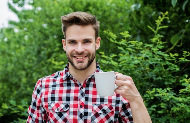 より多くのコーヒーハンサムなひげを生やした男が屋外でお茶を飲む彼はココア料理を愛し、市松模様のシャツで陽気な男を飲む朝のコーヒーおはよう新鮮なインスピレーションとエネルギッシュな飲み物を飲む