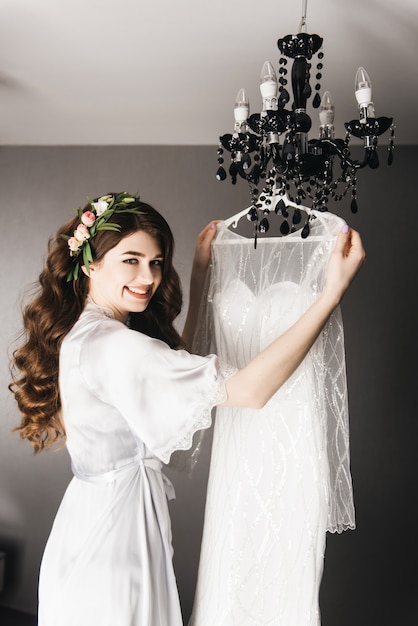 Morning of the bride. Beautiful portrait of a bride in a peignoir with hair curls and fresh flowers near the wedding dress.