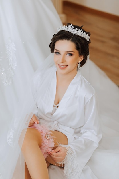 Morning of the bride before the wedding A beautiful young woman with a veil and a beautiful hairstyle in a white robe near her wedding dress on a mannequin Professional makeup Last preparations