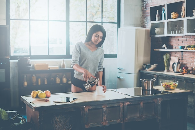 Morning coffee. Beautiful young African woman pouring coffee to cup and smiling while standing in kitchen at home