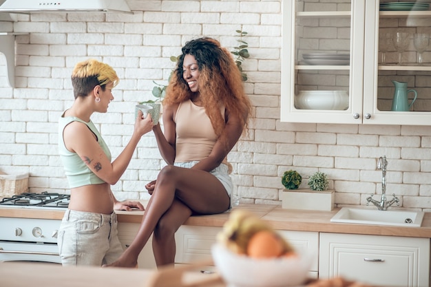 Morning coffee. Dark-skinned joyful woman in shorts barefoot sitting on kitchen table of her girlfriend in jeans standing next to coffee