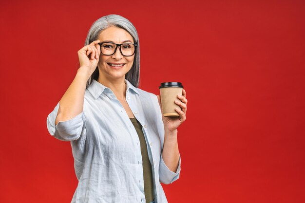 朝のコーヒーに行くフォーマルな服装で魅力的な年配の女性が赤い背景で隔離の熱い飲み物の紙コップで立って良い仕事の日に準備する成熟したビジネスの女性はコーヒー休憩を取る