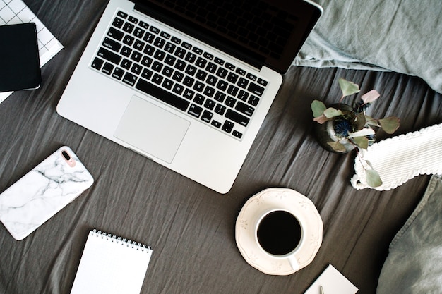 Morning coffee, laptop, mobile with marble case, diary, glasses in bed with grey sheet and pillows