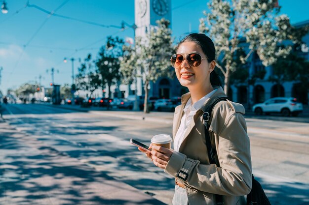 Morning coffee take away lifestyle concept. beautiful young\
woman hold tea paper cup while walking on street with ferry\
building in back. cute smiling lady enjoy sunny day using mobile\
phone outdoor