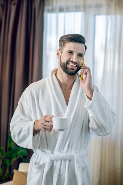 Morning coffee. Young bearded man in a white bath robe having morning coffee and talking on the phone