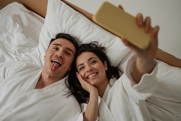 Morning of happy young couple in white bathrobes in bed at home, making selfie.