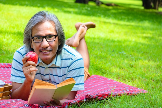 Photo in the morning he is reading book with   red apply.he is  lying on the grass beside picnic