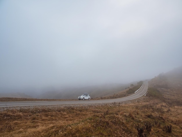 Morning mountain highway in dense fog White car SUV with fog lights turned on is parked next to a scenic route road in a foggy autumn landscape Luggage trunk box mountain on car roof rack