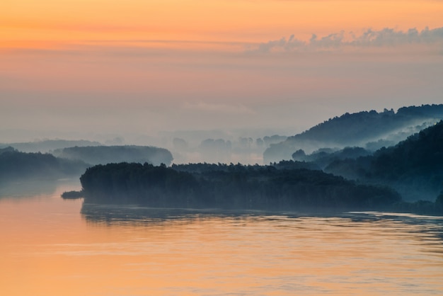 Morning mystic haze boven brede vallei van rivier. Gouden gloed van dageraad in hemel. Rivieroever met bos onder mist. Zonlicht weerspiegeld in water bij zonsopgang. Kleurrijk sfeervol landschap van majestueuze natuur.