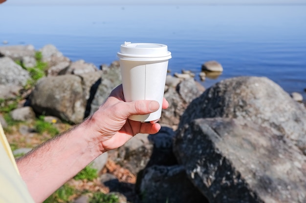 Morning paper coffee cup with tasty hot coffee in hand outdoors on sea wall.Hand holds a white cardboard paper mug for takeaway drinks, against beautiful sea shore wall, environment