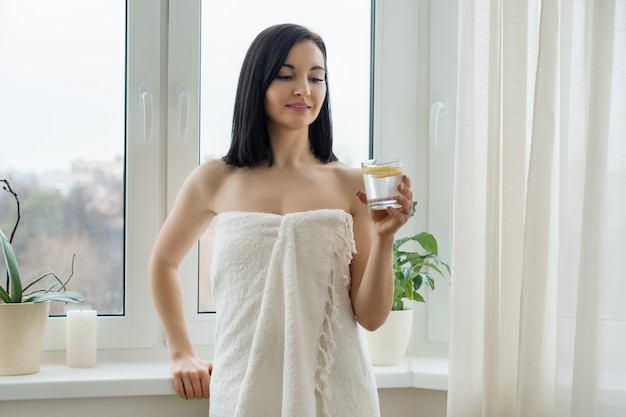 Morning portrait of young beautiful woman in bath towel with glass of water with lemon near the window.