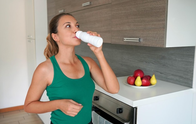 Ritratto mattutino di giovane donna che beve bevande proteiche del siero di latte la ragazza di fitness in casa beve kefir dalla bottiglia